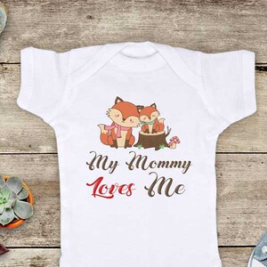 May include: White baby onesie with a graphic of two foxes on a tree stump. The text "My Mommy Loves Me" is printed in red below the foxes.