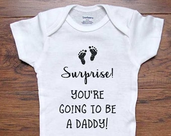 May include: White baby onesie with black text that reads "Surprise! You're going to be a daddy!" and two black footprints above the text.