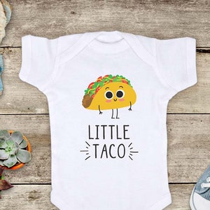 May include: White baby onesie with a cartoon taco graphic and the text "LITTLE TACO". The taco is yellow with green, red, and brown details. The onesie is surrounded by baby toys on a wooden surface.