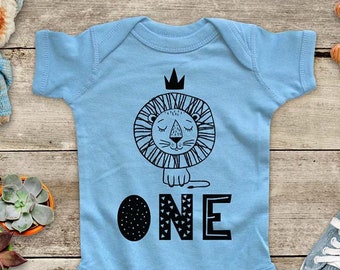 May include: Light blue baby onesie with a black and white lion wearing a crown and the word "ONE" in black and white with a geometric pattern.
