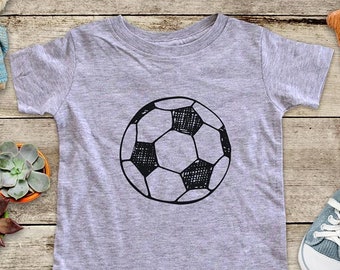 May include: A gray t-shirt with a black and white illustration of a soccer ball. The shirt is on a wooden surface with a variety of decorative items.