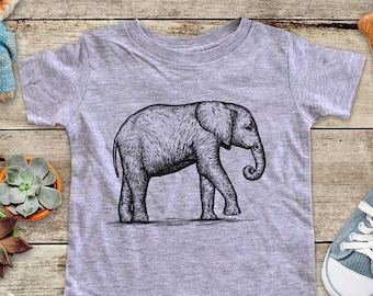 May include: A gray short-sleeved t-shirt featuring a detailed black and white illustration of an elephant. The elephant is depicted walking, with its trunk curled. The shirt is surrounded by children's toys.