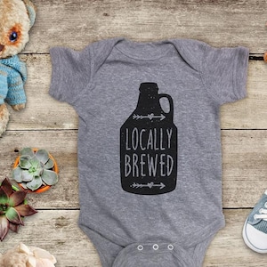 May include: A gray baby onesie with the words "LOCALLY BREWED" printed in black. The onesie features a black graphic of a jug with arrows and hearts. The onesie is surrounded by baby toys.