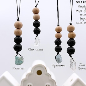 May include: Four hanging ornaments featuring wood beads, black lava rocks, and raw gemstones on black leather cords. Text reads: WOOD & LAVA ROCKS WITH GEMSTONES ON A LEATHER CORD. Simply add a few drops of your favorite essential oil or fragrance.