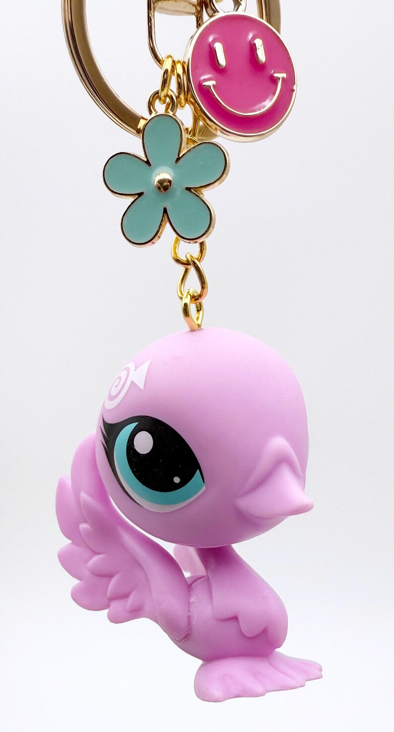 Puede incluir: Un llavero con una figura rosa con forma de pato, grandes ojos azules y un dise&ntilde;o de remolino blanco. El llavero tambi&eacute;n incluye un dije de flor turquesa y un dije de carita sonriente rosa, todo conectado por un anillo dorado.