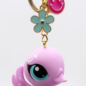 Puede incluir: Un llavero caprichoso con una figura de p&aacute;jaro lavanda con grandes ojos azules y un llavero dorado. El llavero tambi&eacute;n incluye un dije de carita sonriente rosa y un dije de flor turquesa, todo suspendido de un anillo de metal dorado.