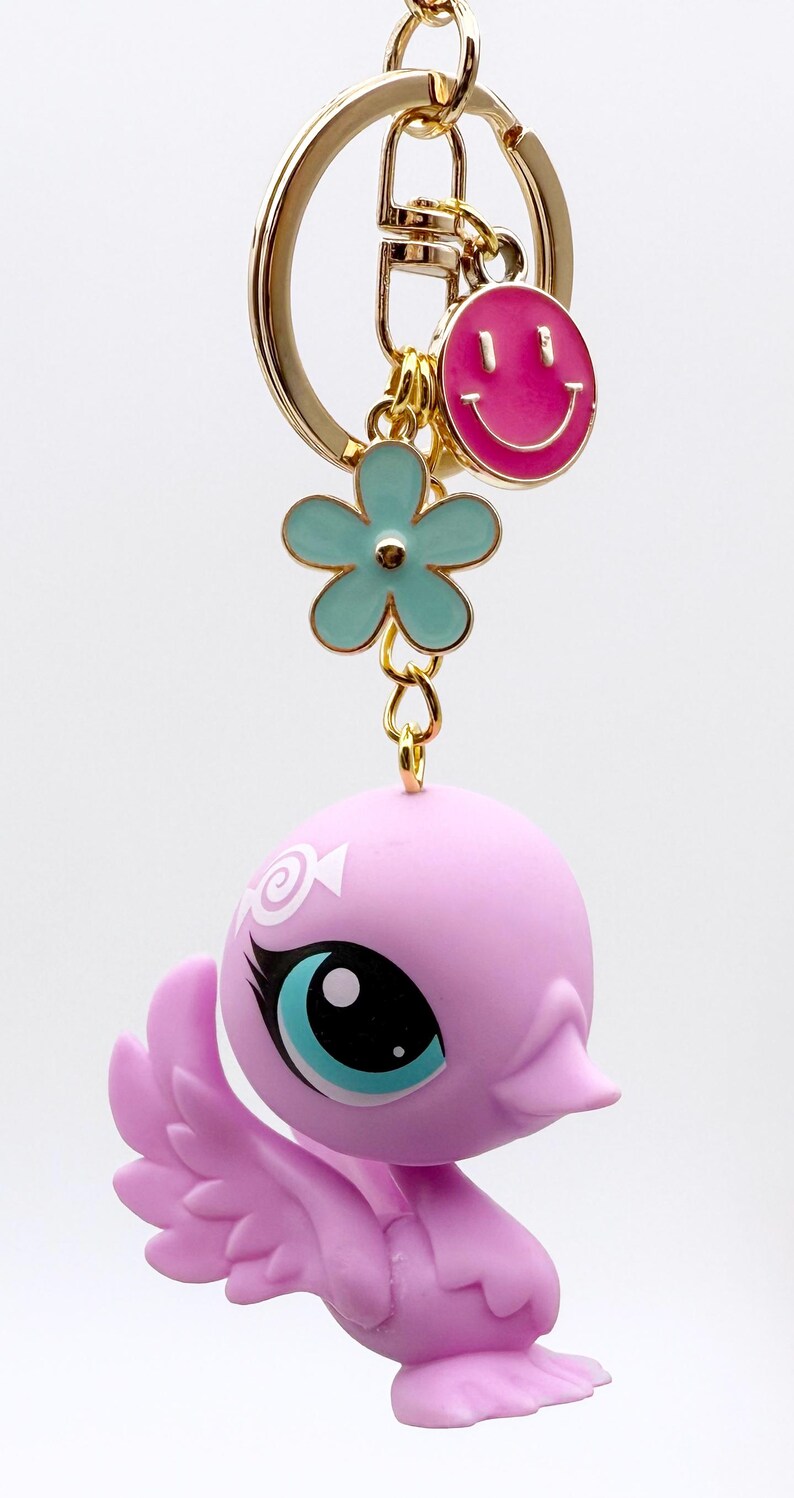 Puede incluir: Un llavero caprichoso con una figura de p&aacute;jaro lavanda con grandes ojos azules y un llavero dorado. El llavero tambi&eacute;n incluye un dije de carita sonriente rosa y un dije de flor turquesa, todo suspendido de un anillo de metal dorado.