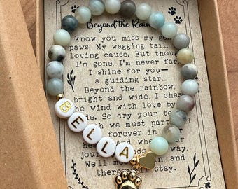 May include: A beaded bracelet featuring smooth, multi-colored stone beads in shades of blue, gray, and cream, white letter beads spelling BELLA in gold, a small gold heart charm, and a gold paw print charm, resting on a card with printed text.
