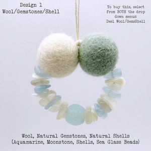 May include: A handcrafted ornament featuring two wool spheres, one cream and one sage green, connected by a string. A ring of aquamarine, moonstone, shells, and sea glass beads forms a decorative element. Text reads "Design 1 Wool/Gemstones/Shell".