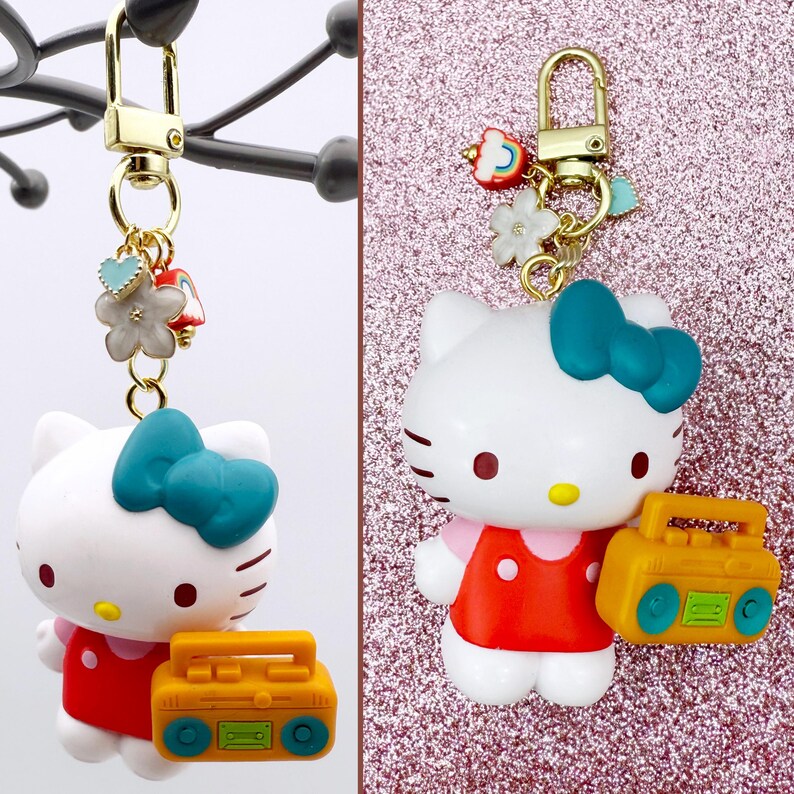 Puede incluir: Un llavero de Hello Kitty blanco con un lazo turquesa, un vestido rojo y un radiocasete amarillo. El llavero tiene un cierre dorado y dijes decorativos, incluyendo flores y un coraz&oacute;n. El fondo es rosa brillante.