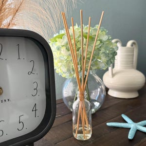 May include: A clear glass aromatherapy diffuser bottle with wooden reeds. A black clock with white numbers is on the left. A vase of green flowers and a white vase are in the background. A blue starfish is on the right.
