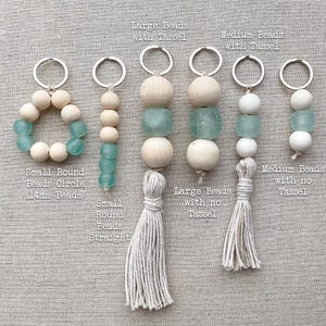 May include: Seven keychains with different bead sizes and styles. Each keychain has a wooden bead, a turquoise glass bead, and a white tassel. The keychains are labeled with the bead size and style, such as "Large Beads with Tassel" and "Small Round Beads Circle (14mm Beads)".