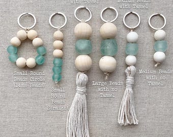 May include: Seven keychains with different bead sizes and styles. Each keychain has a wooden bead, a turquoise glass bead, and a white tassel. The keychains are labeled with the bead size and style, such as "Large Beads with Tassel" and "Small Round Beads Circle (14mm Beads)".