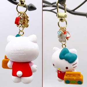 Puede incluir: Un llavero de Hello Kitty blanco con un vestido rojo y un lazo turquesa. El llavero tiene un cierre dorado e incluye dijes de una flor, un coraz&oacute;n y un arco&iacute;ris. La figura sostiene un radiocasete amarillo.