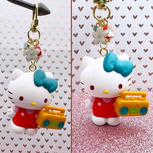 Puede incluir: Un llavero de Hello Kitty blanco con un lazo turquesa, un vestido rojo y un radiocasete amarillo. El llavero tiene un cierre dorado y abalorios decorativos, incluyendo una flor blanca y un peque&ntilde;o objeto rojo. El fondo presenta un patr&oacute;n de corazones.