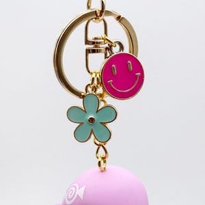 Puede incluir: Un llavero dorado con un charm de carita sonriente rosa, un charm de flor azul claro y una figura rosa con forma de p&aacute;jaro. El llavero tiene un cierre dorado y un gran anillo dorado.