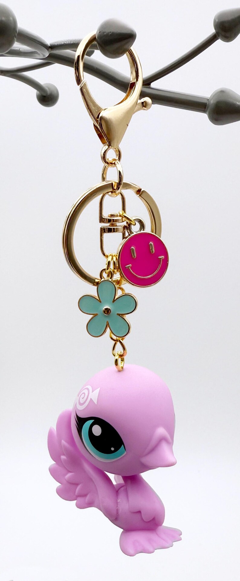 Puede incluir: Un llavero dorado con un charm de carita sonriente rosa, un charm de flor azul claro y una figura rosa con forma de p&aacute;jaro. El llavero tiene un cierre dorado y un gran anillo dorado.