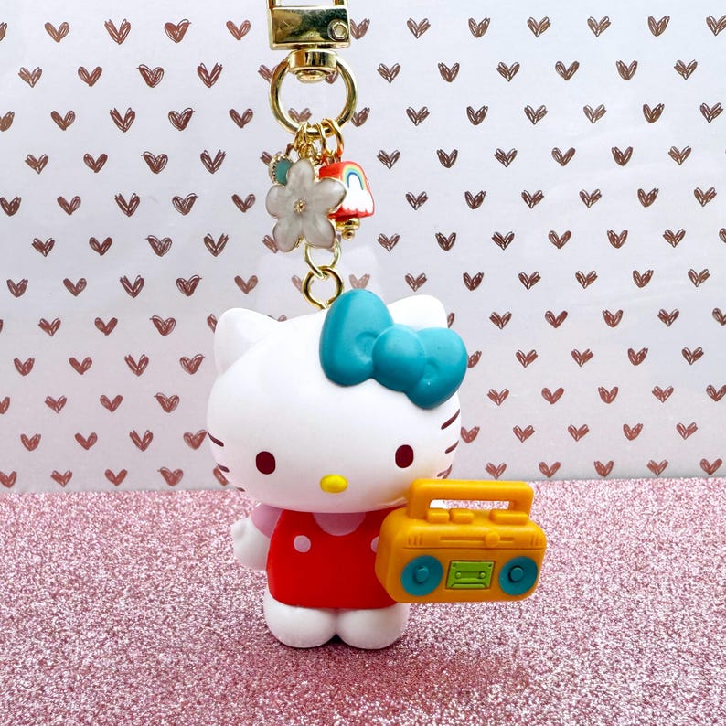 Puede incluir: Un llavero de Hello Kitty blanco con un lazo azul, un vestido rojo y un radiocasete amarillo. El llavero tiene un cierre dorado y abalorios decorativos, sobre una superficie de purpurina rosa y un fondo con estampado de corazones.