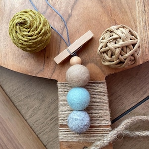 May include: A wooden bead garland with three felt balls in beige, light blue, and gray. The garland is accented with a wooden clothespin and two decorative woven spheres in green and tan. The items are arranged on a wooden surface.