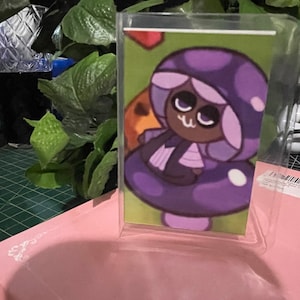 Puede incluir: Una vitrina de acrílico transparente con una imagen impresa de un personaje de dibujos animados. El personaje es una galleta con forma de hongo con un sombrero morado y una cara blanca. El personaje lleva un vestido morado y tiene una cara marrón.