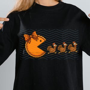 May include: A black short-sleeved t-shirt featuring a graphic of an orange pie shaped like a video game character with a plaid bow, chasing three brown turkeys. The background of the graphic consists of horizontal rows of dark grey chevron lines.