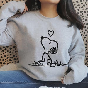 May include: A light gray crewneck sweatshirt featuring a black line art graphic of a cartoon dog sitting on the ground with a small heart shape floating above its head. The sweatshirt has long sleeves and a ribbed neckline, cuffs, and hem.