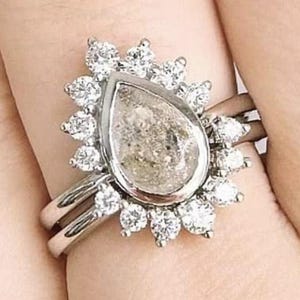 Può includere: Un set di anelli in argento con una pietra centrale a forma di pera, di colore chiaro, circondata da un alone di pietre rotonde e trasparenti più piccole. L'anello è abbinato a una fascia semplice. Il design è elegante e accattivante.