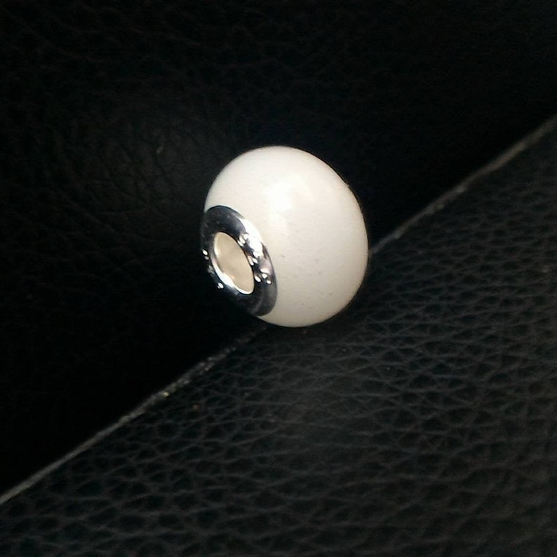 May include: White ceramic bead with a silver metal band around the center. The bead is smooth and glossy.
