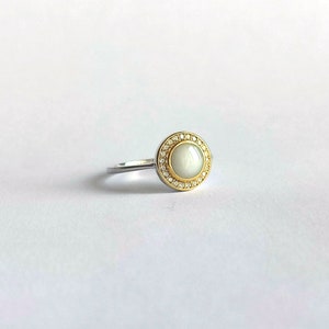 May include: A silver ring with a gold-coloured bezel. The ring features a large, round, off-white stone surrounded by small, clear gemstones. The ring is set against a white background.