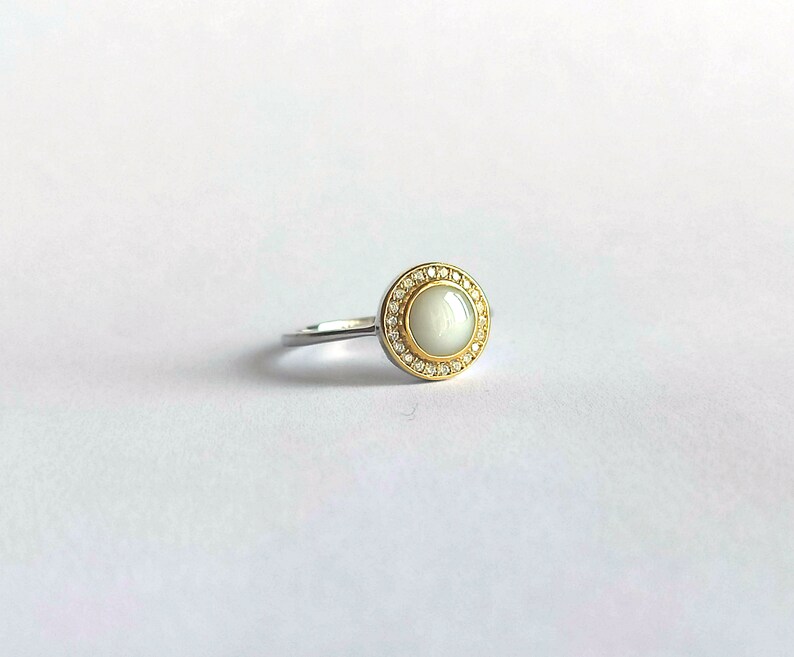 May include: A silver ring with a gold-coloured bezel. The ring features a large, round, off-white stone surrounded by small, clear gemstones. The ring is set against a white background.