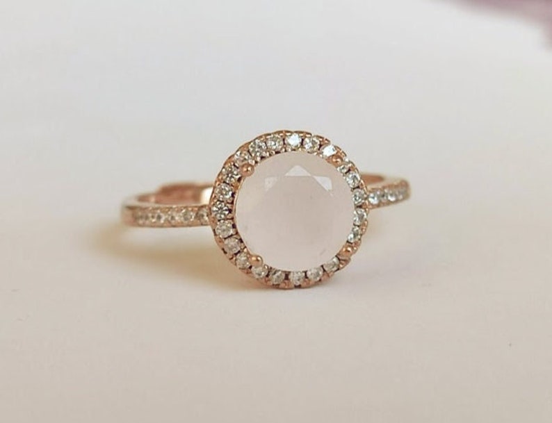 May include: A rose gold ring featuring a round, pale pink gemstone encircled by a halo of small, clear gemstones.
