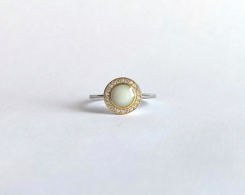 May include: A silver ring featuring a circular, off-white central stone. The stone is encircled by a halo of small, clear gemstones set in a gold-coloured metal. The band is silver-coloured.