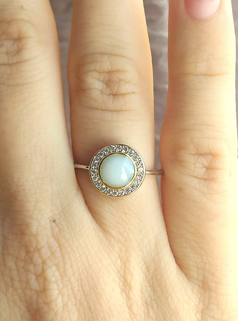 May include: A close-up of a ring featuring a pale blue gemstone surrounded by a halo of small, clear gemstones. The ring has a gold band. The ring is on a finger.