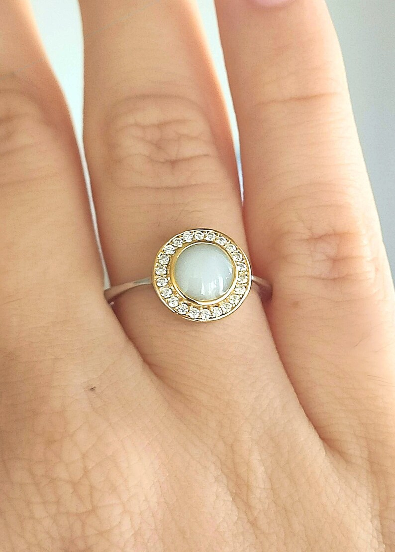 May include: A silver ring featuring a gold-coloured halo set with small clear stones around a pale blue, round gemstone. The ring is on a person's finger.