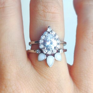 May include: A silver ring with a pear-shaped clear stone surrounded by smaller clear stones. The ring has a band with three white pear-shaped stones.