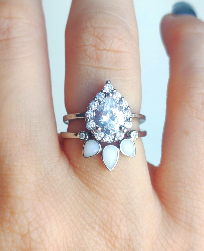 May include: A silver ring with a pear-shaped clear stone surrounded by smaller clear stones. The ring has a band with three white pear-shaped stones.