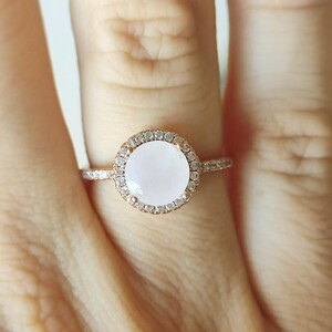 May include: A rose gold ring featuring a large oval-shaped white gemstone set within a halo of smaller clear gemstones.