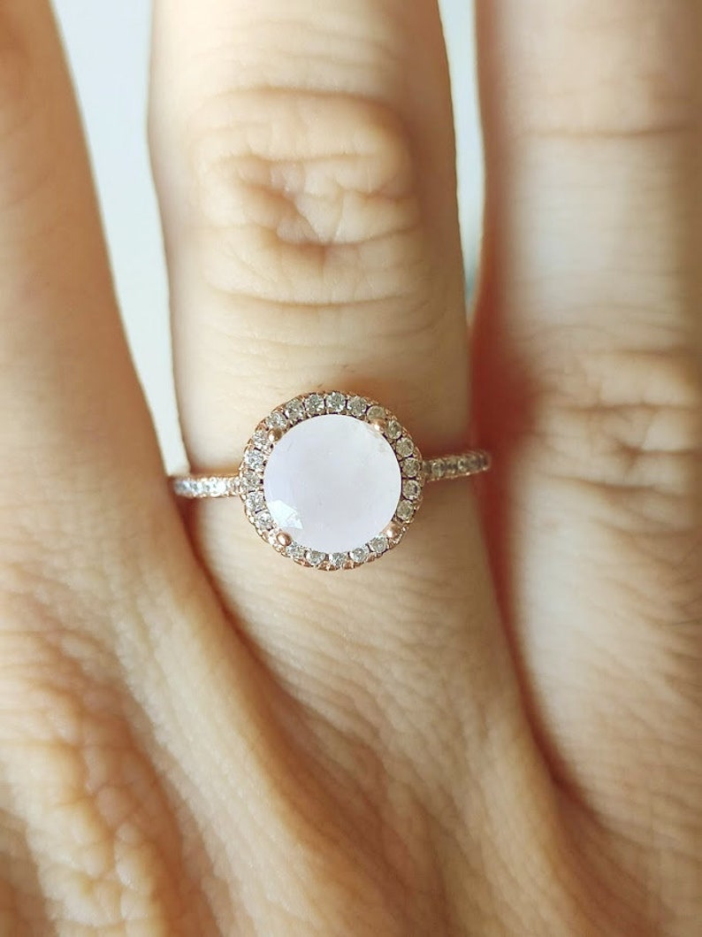 May include: A rose gold ring featuring a large oval-shaped white gemstone set within a halo of smaller clear gemstones.