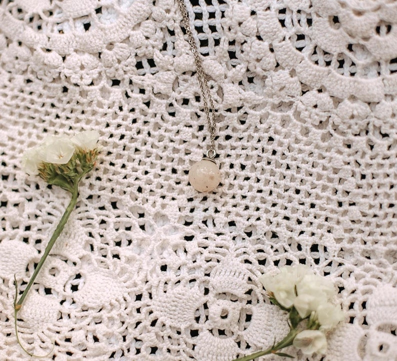 May include: A delicate silver chain necklace with a small, round, light pink gemstone pendant. The necklace is lying on a white crocheted surface with white flowers.