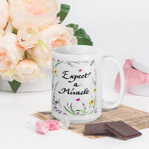 May include: White ceramic mug with a floral design and the text "Expect a Miracle" in black cursive lettering with pink hearts.