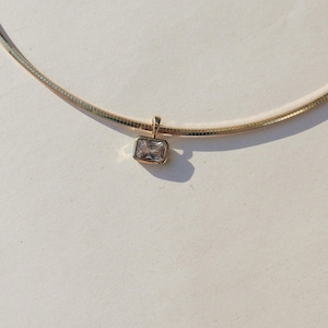 Può includere: Una collana dorata con un piccolo pendente rettangolare. Il pendente presenta una pietra trasparente a taglio quadrato. La collana ha una catena liscia e flessibile. Il gioiello è esposto su una superficie bianca.