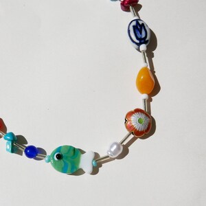 May include: A colorful beaded necklace featuring a green and white fish charm, blue and white patterned beads, and a flower-shaped bead with red and orange accents. The necklace also includes a variety of other colorful beads.