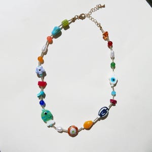 May include: A colorful beaded necklace featuring a variety of shapes and colors. The necklace includes beads in shades of blue, green, orange, red, and white, with a gold-colored chain and clasp. The beads include a fish, flower, and evil eye charm.