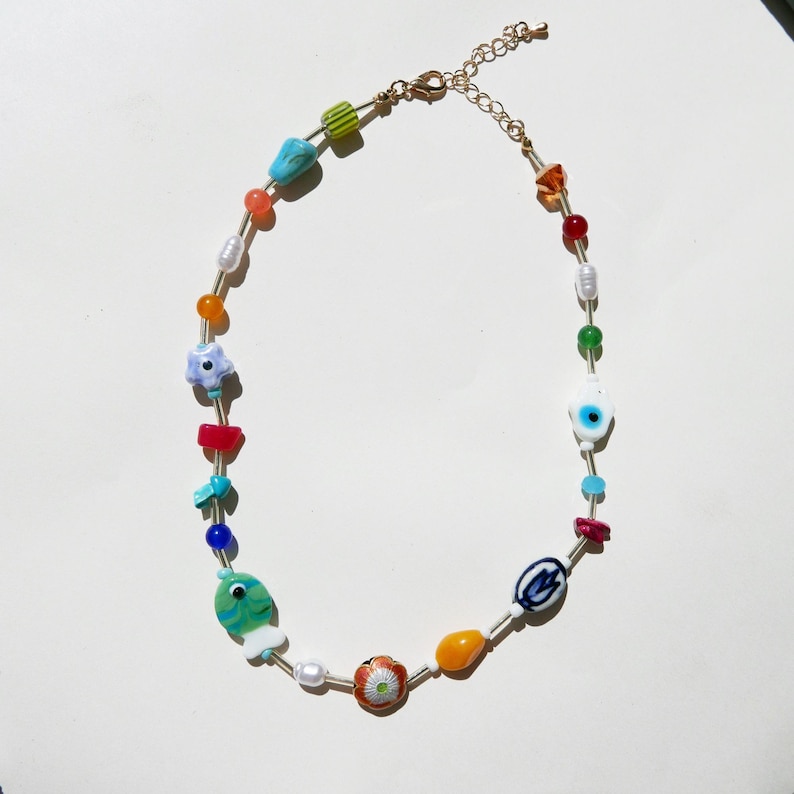 May include: A colorful beaded necklace featuring a variety of shapes and colors. The necklace includes beads in shades of blue, green, orange, red, and white, with a gold-colored chain and clasp. The beads include a fish, flower, and evil eye charm.