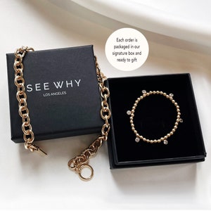 May include: A gold chain necklace and a gold bracelet with small clear stones in a black box. The box has the text "SEE WHY LOS ANGELES" on the top. The text "Each order is packaged in our signature box and ready to gift" is written on a white circle.