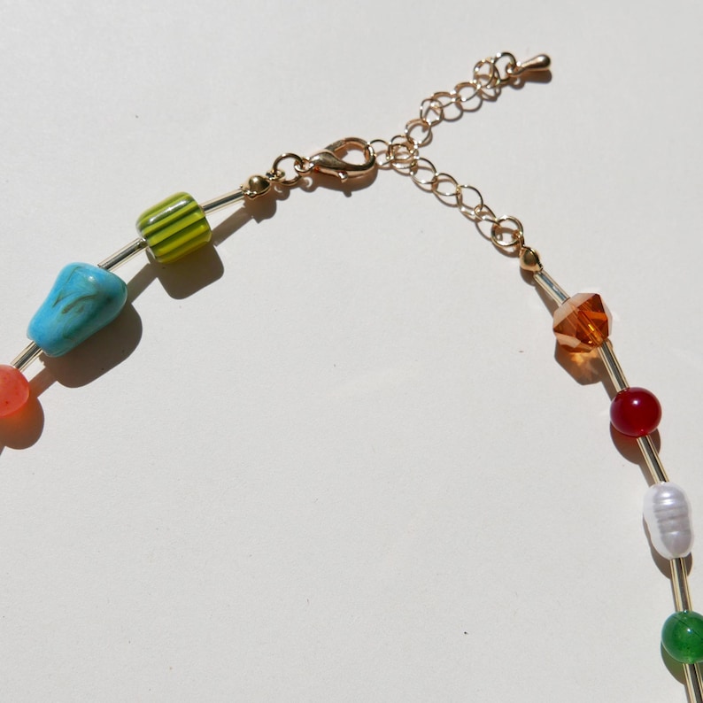 May include: A colorful beaded necklace with a gold-tone chain and clasp. The necklace features a variety of beads in shades of green, blue, orange, red, and white. The beads are strung on a thin wire.