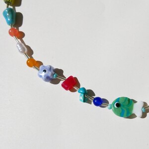 May include: A colorful beaded necklace featuring a variety of shapes and colors. The necklace includes turquoise, orange, red, blue, and green beads, with a fish-shaped pendant at the end. The beads are strung on a silver wire.
