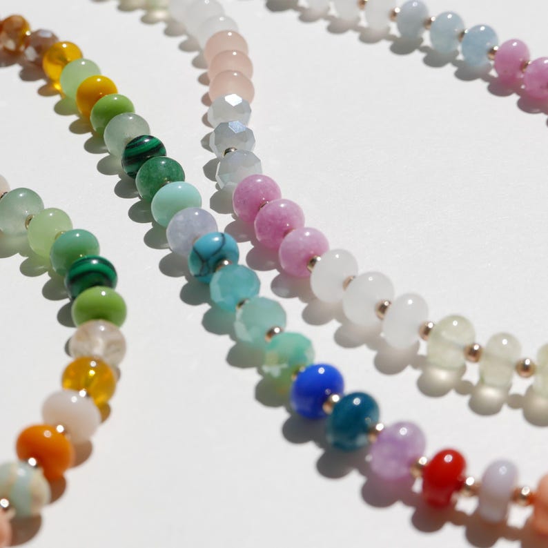 May include: Close-up of several beaded necklaces in a variety of colors. The beads are round and smooth, with a mix of opaque and translucent finishes. The necklaces are arranged on a white surface.