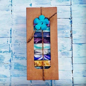 May include: A brown rectangular gift box tied with twine, showcasing a selection of colorful, marbled soaps. The box is adorned with a turquoise flower. The soaps feature shades of blue, purple, white, and gold, creating a visually appealing presentation.