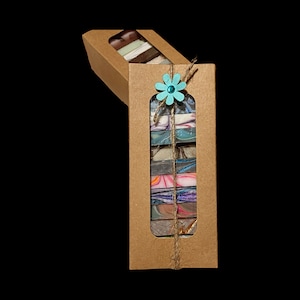 May include: A brown cardboard box with a window showing a stack of colorful handmade soap bars. The box is tied with twine and decorated with a blue flower.